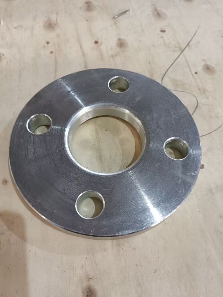 This image shows a metal slip-on flange: circular, with machined metallic texture, a central through-hole, and multiple bolt holes around the edge. It’s placed on a light wooden surface, giving an industrial fitting look.