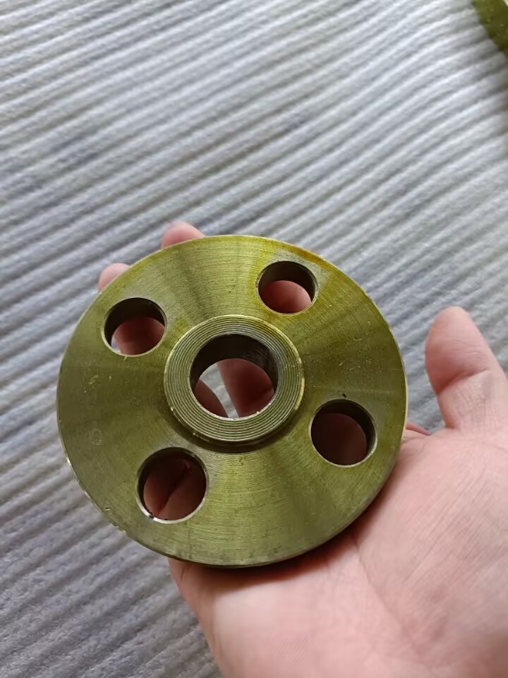 Esta imagen muestra una pequeña brida plana roscada de latón sostenida en la mano. Presenta una superficie de latón dorado con finas texturas mecanizadas, un orificio central con rosca interna y cuatro orificios de montaje distribuidos uniformemente a su alrededor. La brida, colocada sobre un fondo gris texturizado, presenta una apariencia compacta y de precisión: es un accesorio para conexiones de tuberías de diámetro pequeño.