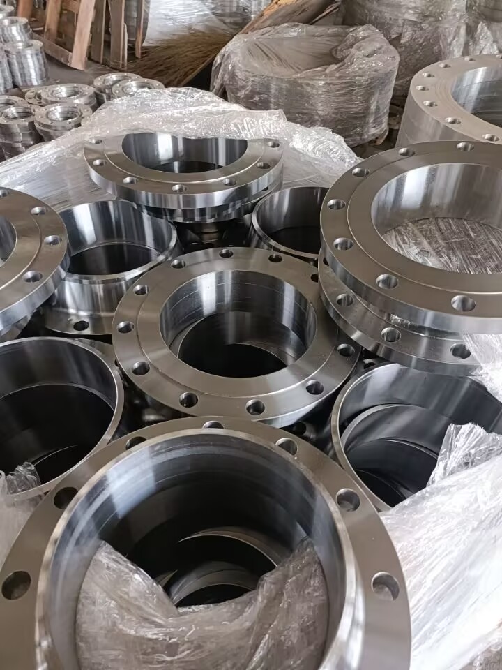 これらは、工場に積み上げられた研磨された金属製の溶接ネックフランジ（おそらくステンレス鋼または炭素鋼製）です。ボルト穴と短いネックがあり、一部はプラスチックで覆われています。典型的な工業用配管継手です。