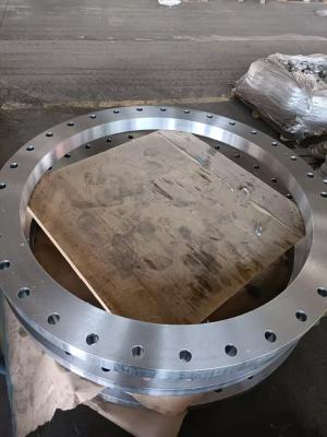 This image displays a large, heavy-duty stainless steel flat flange, situated in a workshop environment. It features a polished metallic surface with numerous evenly spaced mounting holes around its edge, and a wooden board is placed inside the flange (likely for protection). In the background, wrapped batches of similar components are visible. As a large-diameter pipeline fitting, this flange is designed for heavy industrial pipe connections (e.g., in petrochemical or energy systems), providing a secure se